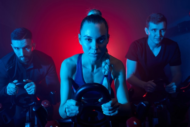 Menschen, die auf dem Heimtrainer im Fitnessstudio trainieren, intensives Cardio-Training. Männer und Frauen trainieren gemeinsam im sportlichen Outfit in einem rauchigen Raum mit blauem Neonlicht