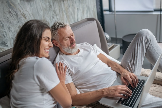 Menschen die einem nahe sind. Konzentrierter grauhaariger Ehemann, der auf Laptoptastatur tippt, und freudige lachende Frau, die Schulter auf Bett berührt