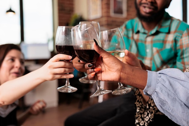 Menschen einer multiethnischen Gruppe überreichen Nahaufnahmen beim Toasten, klirren mit Weingläsern auf einer Wohnungsparty. Freunde verschiedener Nationalitäten feiern, genießen gemeinsam beim Haustreffen.