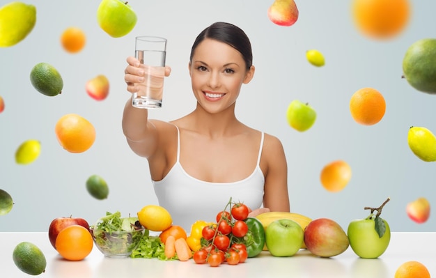 Menschen, Ernährung und vegetarisches Konzept - glückliche Asiatin mit gesundem Essen, die ein Glas Wasser auf grauem Hintergrund mit fallenden Früchten zeigt