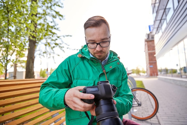 Menschen, Fotografie, Technologie, Freizeit und Lifestyle - junger Hipster-Mann, der eine Digitalkamera mit großem Objektiv auf der Straße der Stadt hält und anschaut