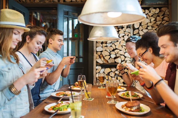 menschen, freizeit, freundschaft, technologie und internetsuchtkonzept - gruppe glücklich lächelnder freunde mit smartphones, die fotos von essen in der bar oder im pub machen