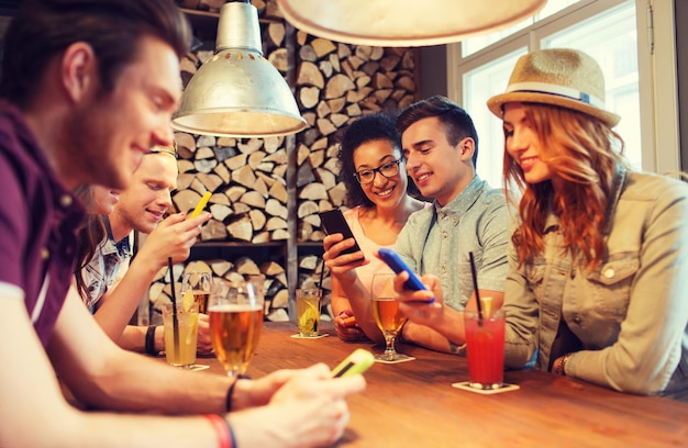 menschen, freizeit, freundschaft, technologie und kommunikationskonzept - gruppe glücklich lächelnder freunde mit smartphones und getränken in der bar oder im pub