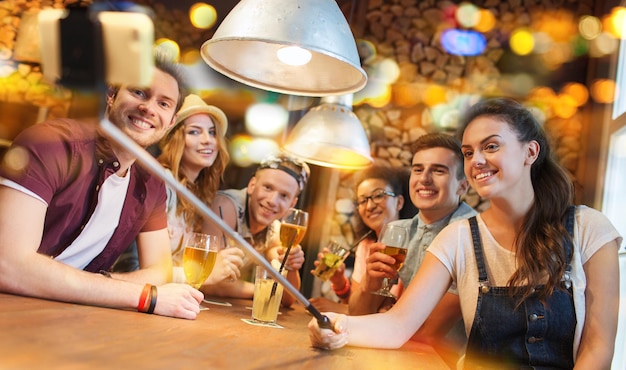 menschen, freizeit, freundschaft, technologie und kommunikationskonzept - gruppe glücklicher lächelnder freunde mit smartphone auf selfie-stick und getränken, die in bar oder pub fotografieren