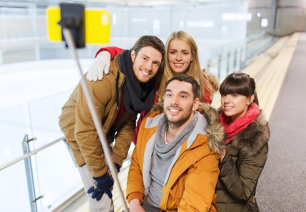 menschen, freundschaft, technologie und freizeitkonzept - glückliche freunde, die mit dem smartphone-selfie-stick auf der eisbahn fotografieren