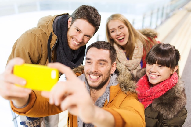 menschen, freundschaft, technologie und freizeitkonzept - glückliche freunde, die sich mit dem smartphone auf der eisbahn selbst machen