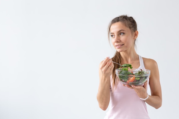 Menschen, gesunder Lebensstil und Fitnesskonzept - Schöne junge Frau nach dem Training, die gesunden Salat auf weißer Wand mit Kopienraum isst
