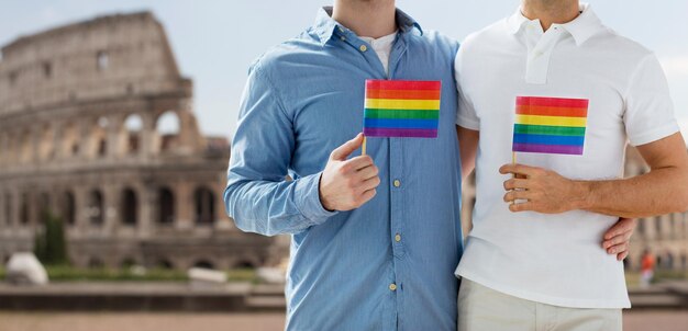 Menschen, Homosexualität, gleichgeschlechtliche Ehe, Reise- und Liebeskonzept - Nahaufnahme eines glücklichen männlichen schwulen Paares, das Regenbogenfahnen hält und sich von hinten über dem Kolosseum im Hintergrund von Rom umarmt