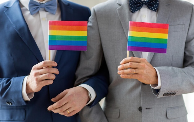 Menschen, Homosexualität, gleichgeschlechtliche Ehe und Liebeskonzept - Nahaufnahme eines glücklichen männlichen schwulen Paares in Anzügen und Fliege mit Eheringen mit Regenbogenfahnen