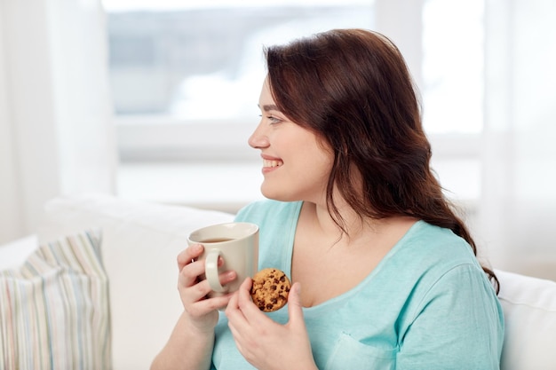 menschen, junk food, ungesunde ernährung und freizeitkonzept - glückliche junge frau in übergröße mit tasse tee mit keks zu hause