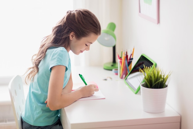 Menschen, Kinder, Bildungs- und Lernkonzept - glückliches Mädchen mit Tablet-PC, das zu Hause in ein Notebook schreibt