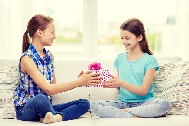 menschen, kinder, ferien, freunde und freundschaftskonzept - glückliche kleine mädchen mit geburtstagsgeschenk sitzen auf dem sofa zu hause