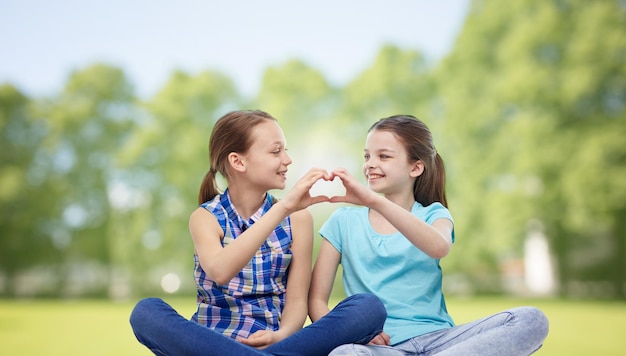 menschen, kinder, freunde und freundschaftskonzept - glückliche kleine mädchen sitzen und zeigen herzförmiges handzeichen über sommerparkhintergrund