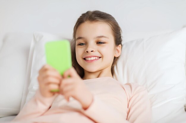menschen, kinder, ruhe und technologiekonzept - glückliches lächelndes mädchen, das zu hause mit smartphone im bett wach liegt