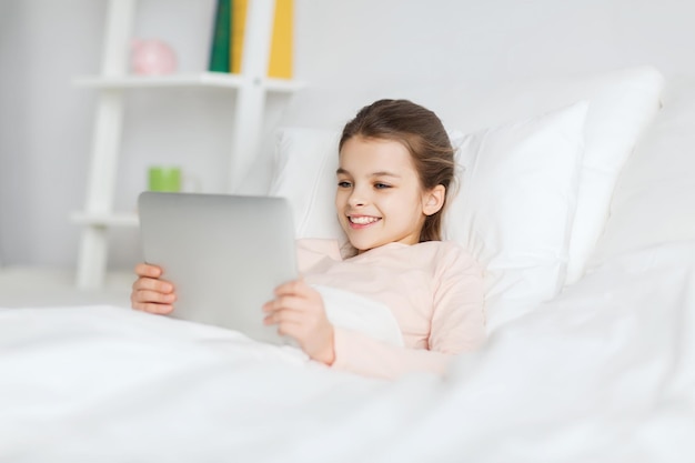 menschen, kinder, ruhe und technologiekonzept - glückliches lächelndes mädchen, das zu hause mit tablet-pc-computer im bett wach liegt