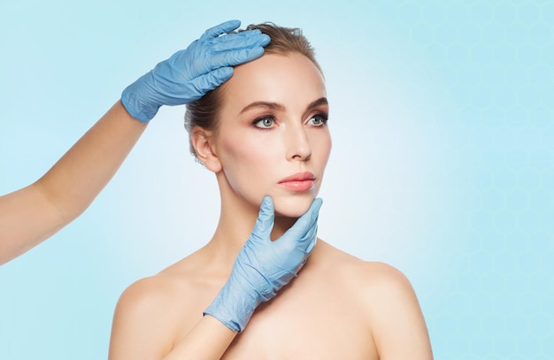 Menschen, Kosmetologie, plastische Chirurgie und Schönheitskonzept - Chirurgen- oder Kosmetikerhände berühren Frauengesicht auf blauem Hintergrund