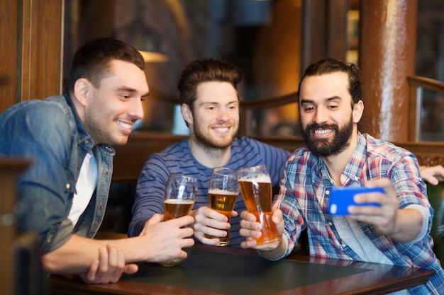 menschen, männer, freizeit, freundschaft und technologiekonzept - glückliche männliche freunde, die bier trinken und selfie mit dem smartphone in der bar oder im pub machen