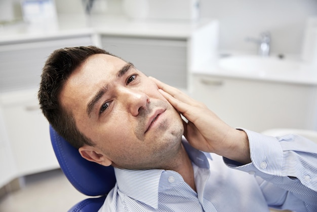 menschen, medizin, stomatologie und gesundheitskonzept - unglücklicher männlicher patient mit zahnschmerzen, der auf einem zahnstuhl im klinikbüro sitzt