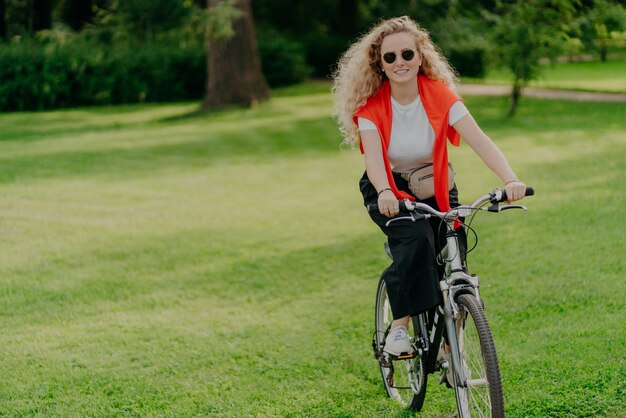 Menschen Natur Rest Lifestyle-Konzept Fröhliche, lockige Frau fährt Fahrrad zwischen grünem Gras, bewegt sich aktiv will fit sein, erkundet neue Orte auf dem Land und trägt Sonnenbrillen, Freizeitkleidung