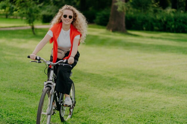 Menschen Natur Rest Lifestyle-Konzept Fröhliche, lockige Frau fährt Fahrrad zwischen grünem Gras, bewegt sich aktiv will fit sein, erkundet neue Orte auf dem Land und trägt Sonnenbrillen, Freizeitkleidung