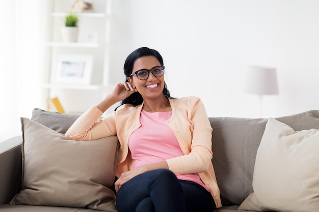 Menschen, Rasse, ethnische Zugehörigkeit und Freizeitkonzept - glücklich lächelnde junge indische Frau mit Brille sitzt zu Hause auf dem Sofa