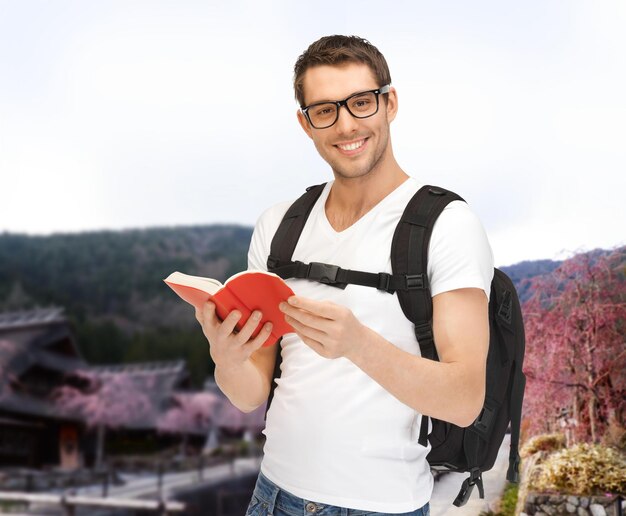 menschen-, reise-, tourismus- und bildungskonzept - glücklicher junger mann mit rucksack und buch, der über asiatischen landschaftshintergrund reist