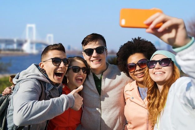 menschen, reisen, tourismus, freundschaft und technologiekonzept - gruppe glücklicher jugendlicher freunde, die selfie mit smartphone machen und daumen hoch über der regenbogenbrücke in tokio im japanischen hintergrund zeigen