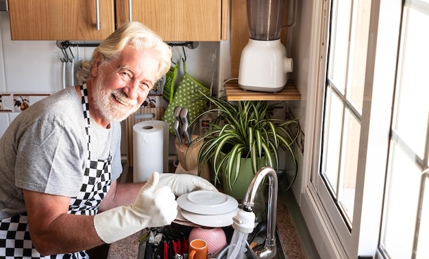 Foto menschen senior mann weiße haare und bart tragen schürze und handschuhe beim abwasch ecke der küche großvater bei der arbeit helles licht aus dem fenster