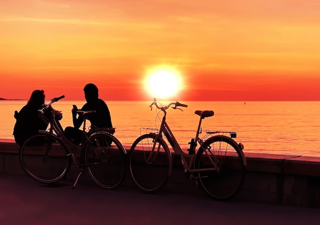 Menschen silhouette romantisches paar mit fahrrad sitzen auf goldorange sonnenuntergang an der seepromenade