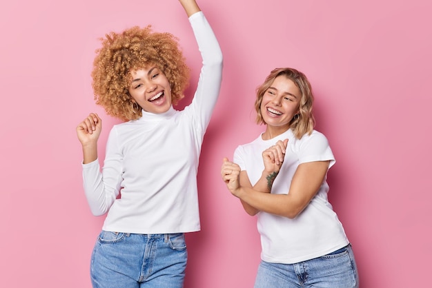 Foto menschen spaß unterhaltungskonzept positive sorgenfreie frauen tanzen und singen zusammen, schütteln die arme in freizeitkleidung, bewegen sich vor rosa hintergrund zwei frohe mädchen bewegen sich zur musik, als würde niemand zuschauen