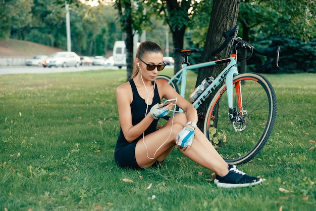 Menschen, Sport und Lifestyle-Konzept - Schönes Mädchen, das in einer warmen Tagesnatur mit einem Fahrrad, einem Telefon und Kopfhörern spaziert, hübsches süßes Mädchen, das an einem sonnigen Tag Smartphone neben ihrem Fahrrad in Parkpalmen benutzt