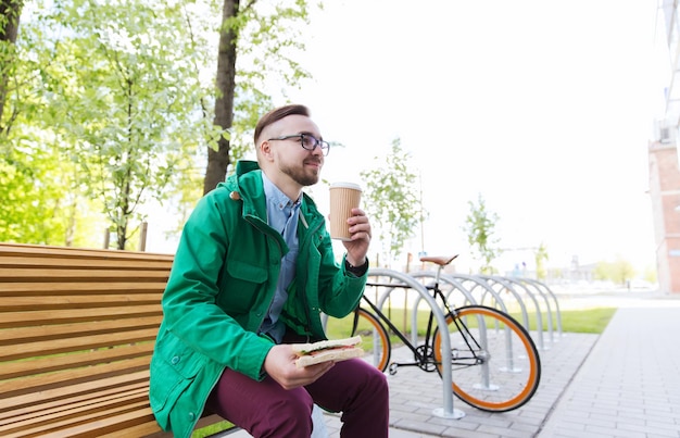 menschen, stil, freizeit und lifestyle - glücklicher junger hipster-mann mit sandwich, kaffeetasse und festradfahrrad, das auf der stadtstraße geparkt ist