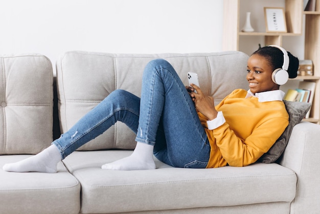 Menschen, Technologie und Freizeitkonzept - glückliche afroamerikanische junge Frau, die mit Smartphone und Kopfhörern auf dem Sofa sitzt und zu Hause Musik hört