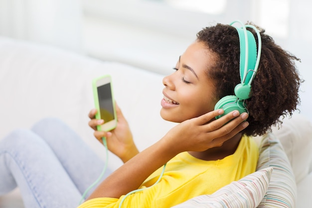 Menschen, Technologie und Freizeitkonzept - glückliche afroamerikanische junge Frau, die mit Smartphone und Kopfhörern auf dem Sofa sitzt und zu Hause Musik hört