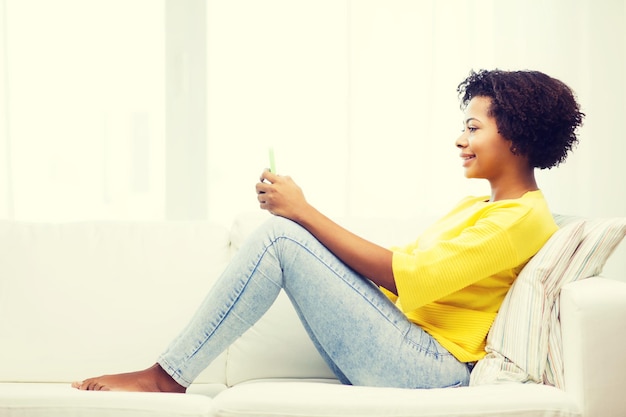menschen-, technologie- und freizeitkonzept - glückliche afroamerikanische junge frau, die zu hause mit smartphone auf sofa sitzt