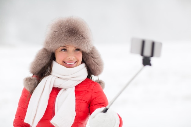 Menschen, Technologie und Freizeitkonzept - glückliche lächelnde Frau in Winterfellmütze fotografiert mit dem Smartphone auf Selfie-Stick im Freien