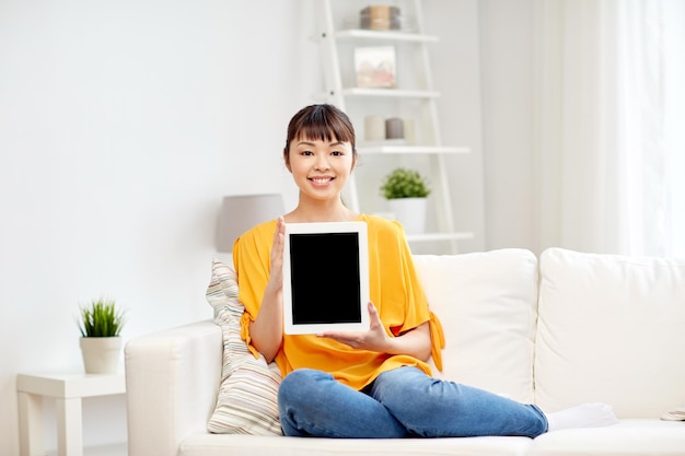 menschen, technologie, werbung und freizeitkonzept - glückliche junge asiatin, die auf dem sofa sitzt und zu hause einen leeren schwarzen bildschirm des tablet-pc-computers zeigt