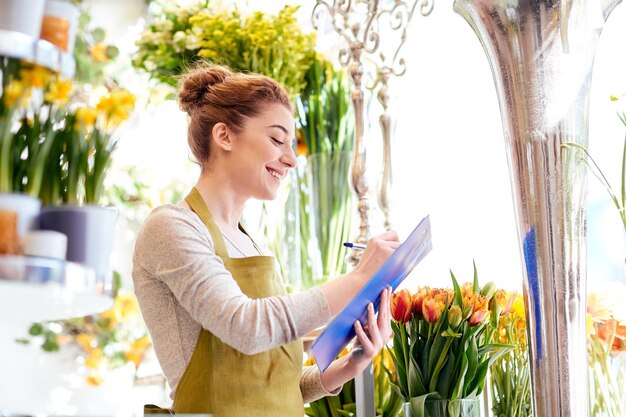 menschen, verkauf, einzelhandel, geschäft und floristikkonzept - glücklich lächelnde floristin mit klemmbrett schreiben und notizen bestellen im blumenladen