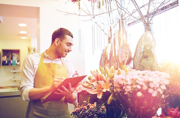 menschen, verkauf, einzelhandel, geschäft und floristikkonzept - glücklich lächelnder floristenmann mit klemmbrett schreiben und notizen bestellen im blumenladen