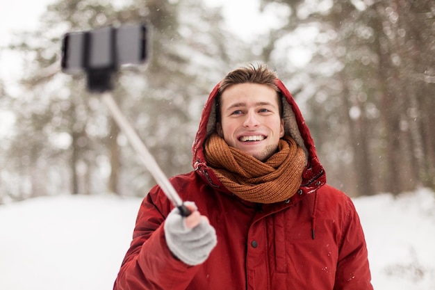 Menschen, Weihnachten, Winter und Jahreszeitenkonzept - glücklich lächelnder Mann mit Kapuze und Schal, der draußen ein Foto mit einem Smartphone-Selfie-Stick macht