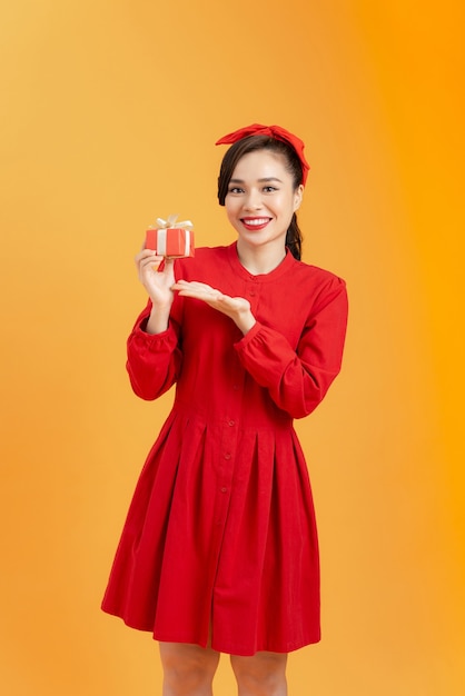 Menschen-, Weihnachts-, Geburtstags- und Feiertagskonzept - glückliche junge Frau im roten Kleid, das mit Geschenkbox über orangefarbenem Hintergrund zeigt/hält.