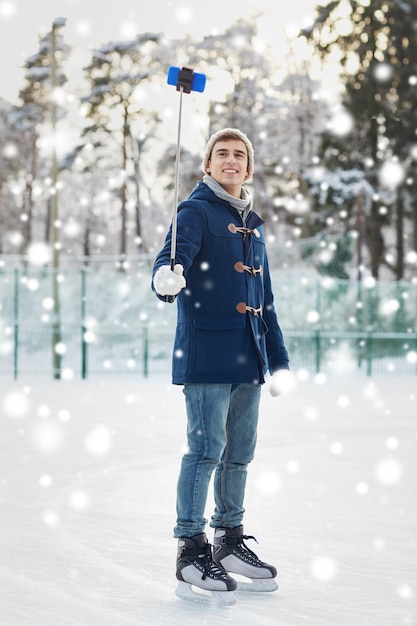 Menschen, Winter, Technologie und Freizeitkonzept - glücklicher junger Mann, der mit Smartphone-Selfie-Stick auf der Eislaufbahn im Freien fotografiert