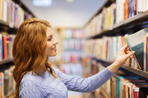 menschen, wissen, bildung und schulkonzept - glückliches studentenmädchen oder junge frau, die buch aus dem regal in der bibliothek nimmt