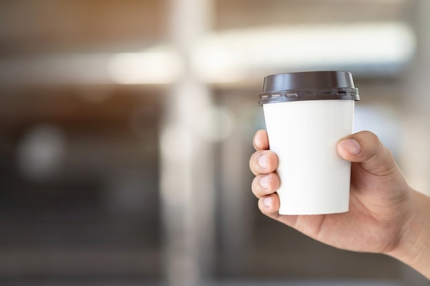 Menschenmannhand, die Pappbecher zum Mitnehmen des Kaffeetrinkens auf natürlichem Morgensonnenlicht hält, das im öffentlichen Park sitzt.