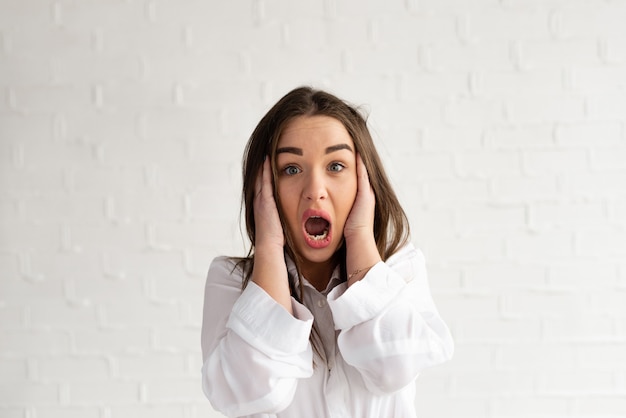 Menschliche Gesichtsausdrücke und Emotionen. Junge erwachsene Frau, die Arme auf ihren Wangen hält und schockiert. Auf weißem Hintergrund isoliert