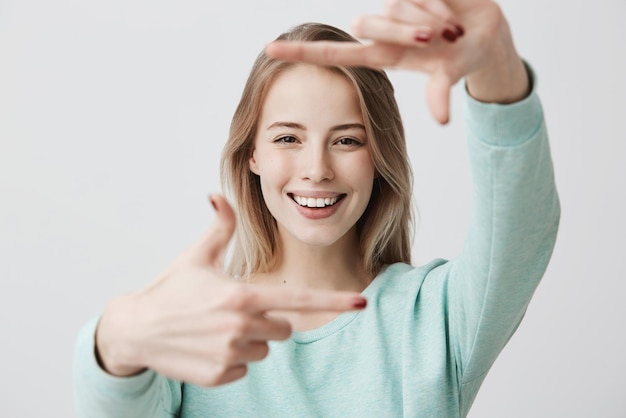 Menschliche Gesichtsausdrücke und Emotionen Positive junge schöne Frau mit gefärbten blonden glatten Haaren in Freizeitkleidung, die mit ihren Fingern ein Quadrat macht, die Kamera anschaut und angenehm lächelt