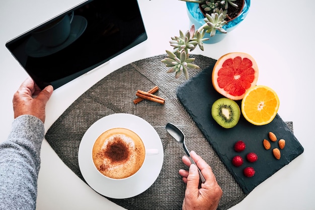 Menschliche Hände halten einen Teelöffel und berühren die Tablette Weiße Tasse hausgemachter Cappuccino-Kaffee und Milchfrüchte für ein gesundes Frühstück