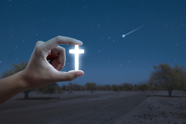 Menschliche Hand, die christliches Kreuz mit dem Nachtszenenhintergrund hält