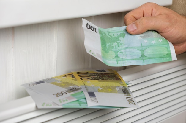 Menschliche Hand, die Euro-Banknoten in der Nähe des elektrischen Heizkörpers zu Hause hält. Mann zahlt Geld für Heizung