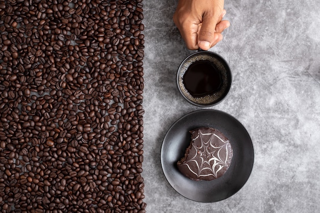 Menschliche Handgriffkaffeetasse und Schokoladenkuchen mit Kaffeebohnen auf Zement masern Hintergrund.